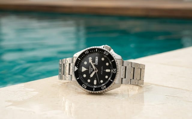 Seiko 5 Sports SRPD55K1 automatic steel watch with black dial by the edge of a turquoise swimming pool, water droplets visible on the crystal