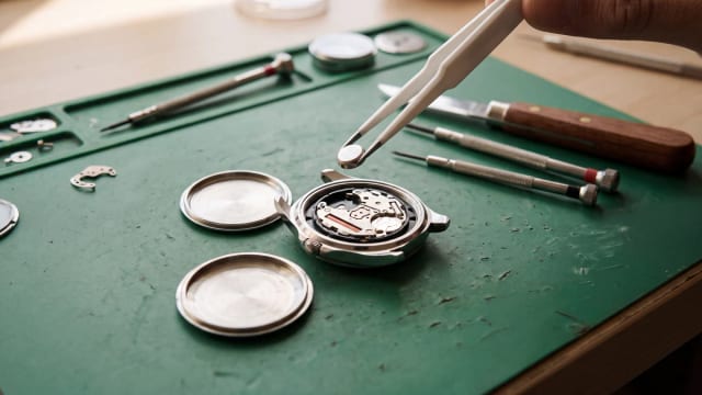 Silver oxide button cell held by plastic tweezers above a quartz movement, watch case back open on a workbench