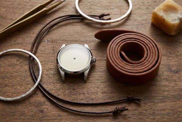 Boîtier de montre en acier, bracelet en cuir et bijoux artisanaux en argent réunis à plat sur un établi en bois