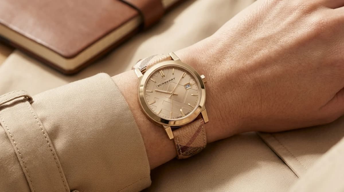 Poignet féminin portant la vraie montre Burberry The City BU9026 argentée sur bracelet cuir beige monogramme, avec trench coat et carnet en arrière-plan