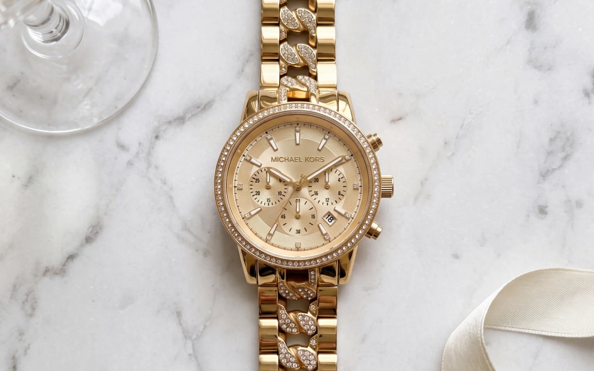 Michael Kors Ritz MK6937 gold-tone chronograph with crystal bezel on marble beside a crystal coupe