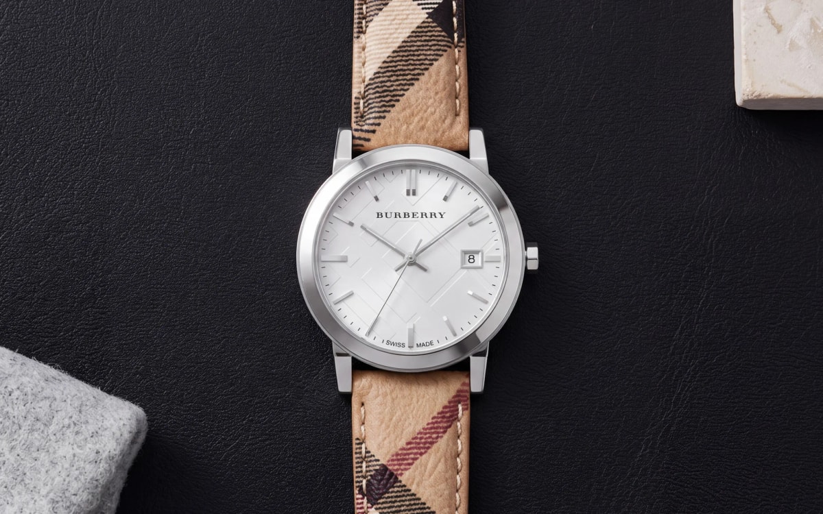 Montre femme Burberry The City BU9025 en acier sur bracelet toile check, posée sur un sous-main en cuir noir
