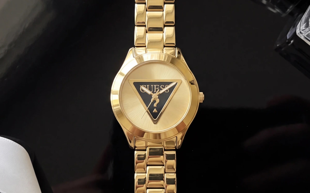 Montre femme GUESS GW0675L2 dorée, cadran au triangle noir, posée sur une laque noire près d'un ruban de satin