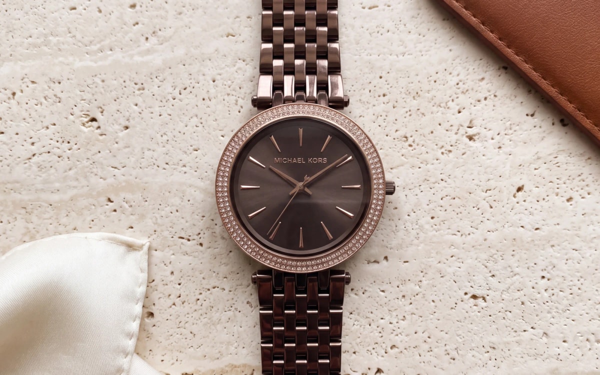 Montre femme Michael Kors Darci MK3416 en acier marron, cadran chocolat et lunette sertie, posée sur travertin