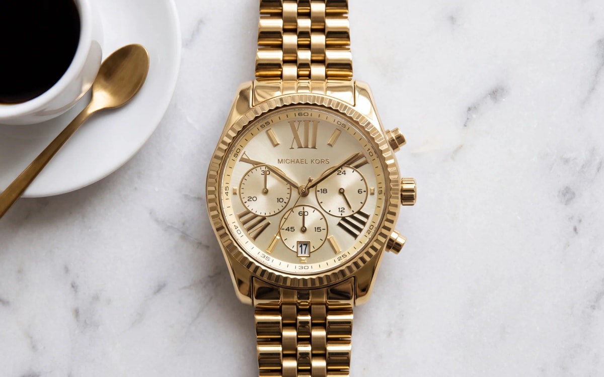 Montre femme Michael Kors Lexington MK5556 dorée, chronographe posé sur marbre blanc près d'un expresso