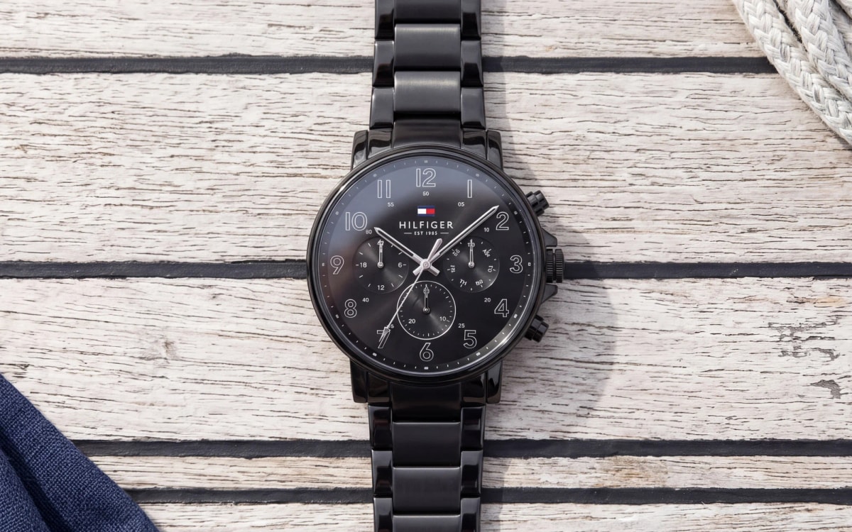 Montre homme Tommy Hilfiger 1710383 noire intégrale, chiffres arabes et trois compteurs, posée sur un pont en teck